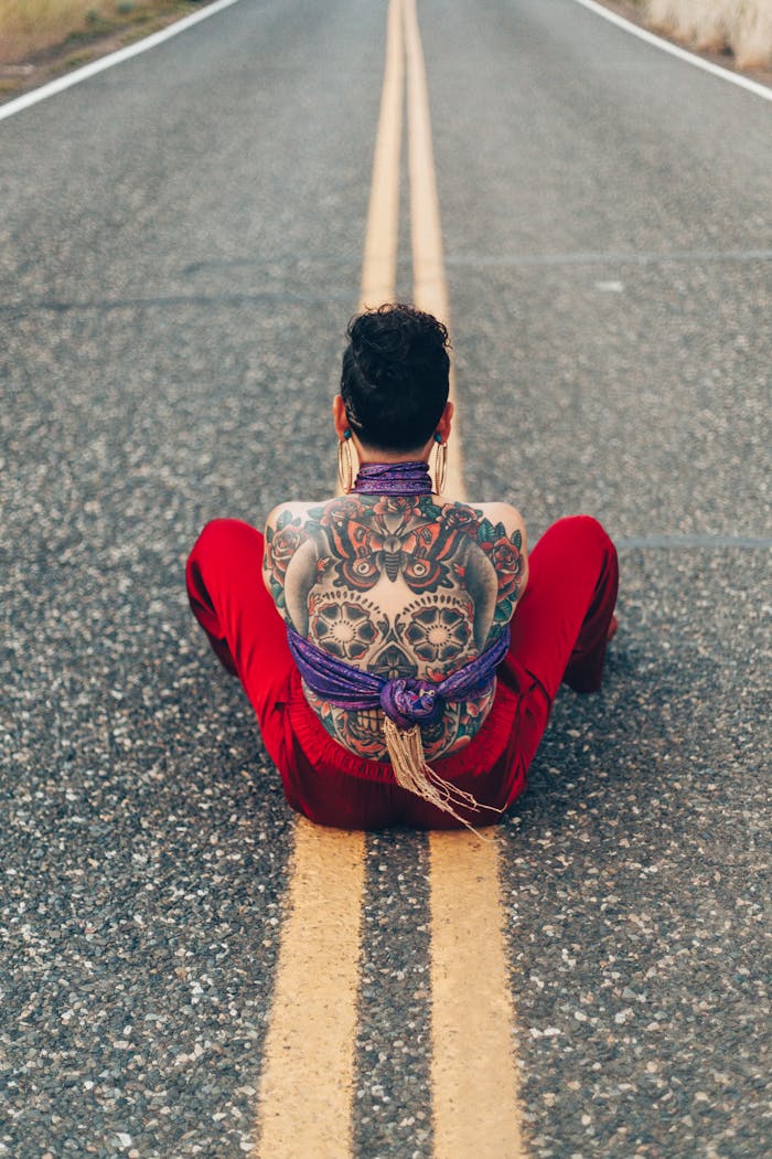 Tattooed person with calavera design sits tied on deserted road, back view.