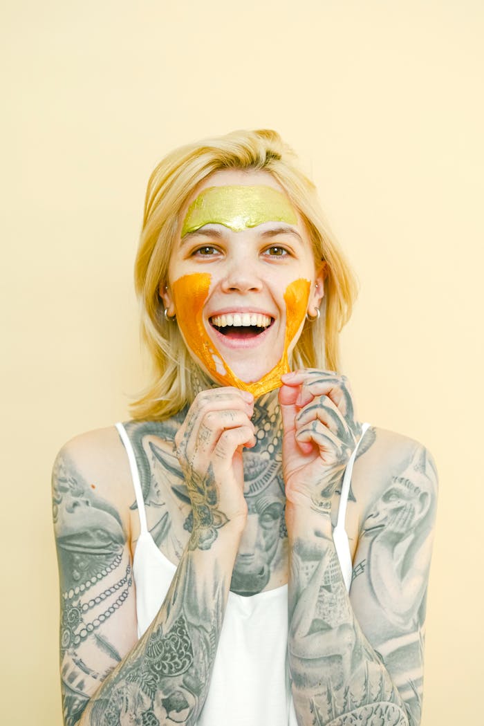 about-02 Young cheerful female with tattooed body and blond hair removing peel off golden mask from face looking at camera