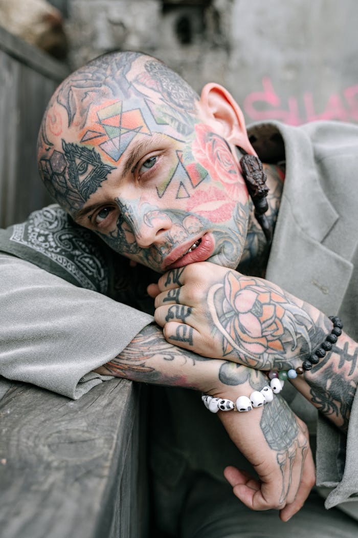 Close-up portrait of a tattooed man in a contemplative pose outdoors, showcasing intricate facial and hand tattoos.