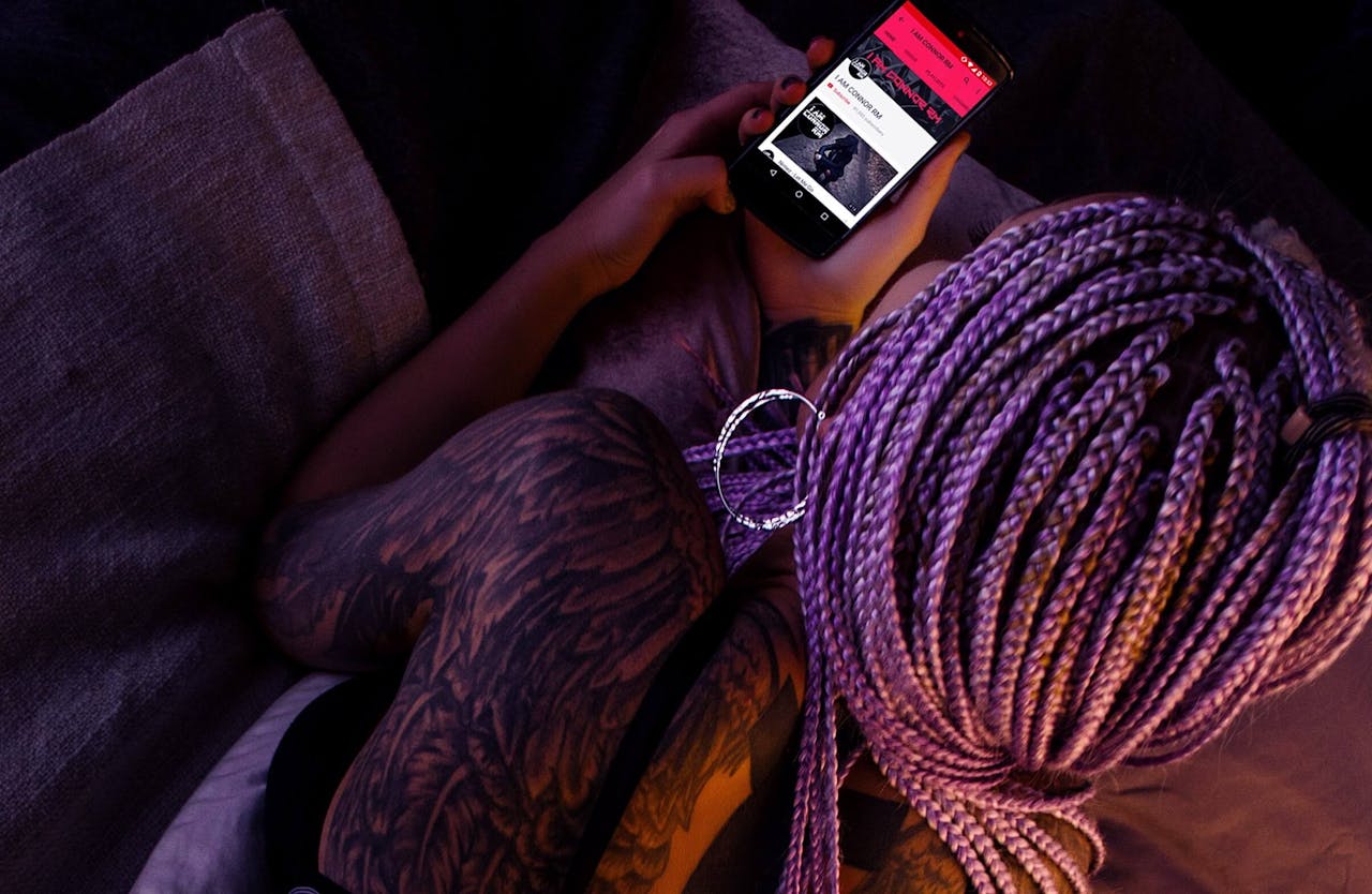 menu-13 A young woman with purple braided hair and tattoos using a smartphone in a dimly lit environment.
